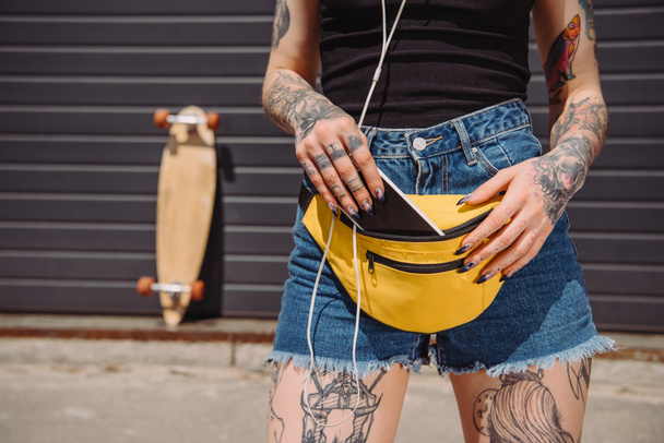 cropped image of tattooed girl putting smartphone with blank screen in waist bag  - Fotó, kép