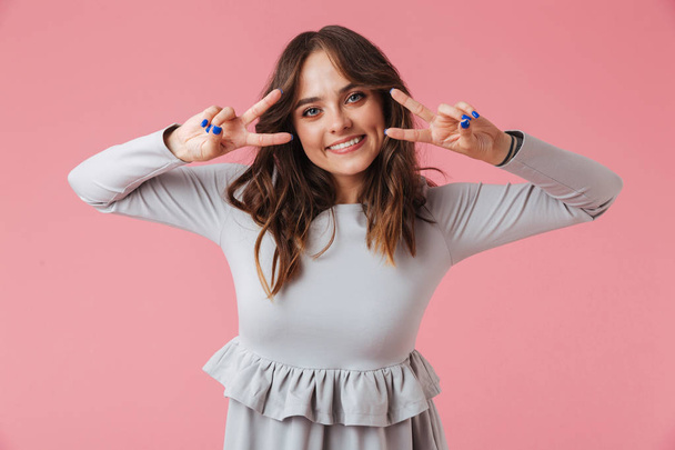 Photo of cheerful young pretty woman standing isolated over pink wall background looking camera make peace gesture. - Photo, image