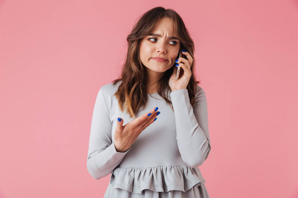 Portrait of a confused young girl talking on mobile phone isolated over pink background - Photo, image