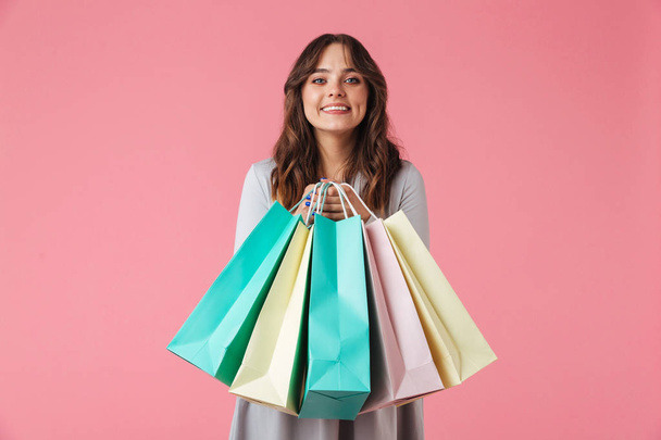 Photo of happy young pretty woman shopaholic standing isolated over pink wall background looking camera holding shopping bags. - Фото, изображение
