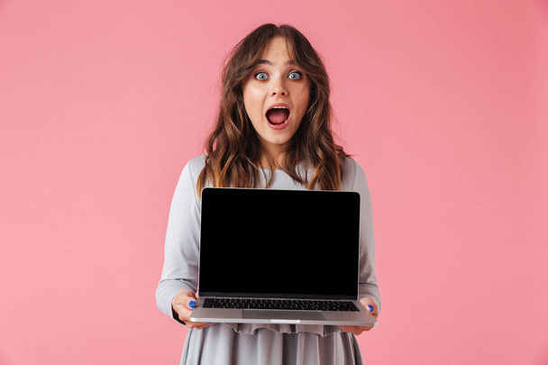 Portrait of an excited young girl showing blank screen laptop computer isolated over pink background - Foto, imagen