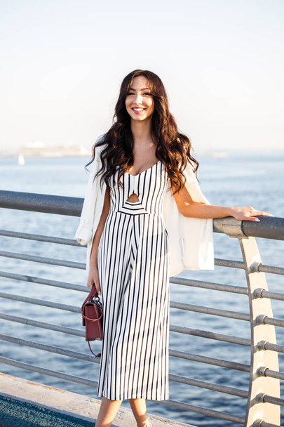 Outdoor shot of beautiful young brunette woman in striped black and white dress. Curly woman standing on pier during sunset, blurred sea background  - Photo, Image