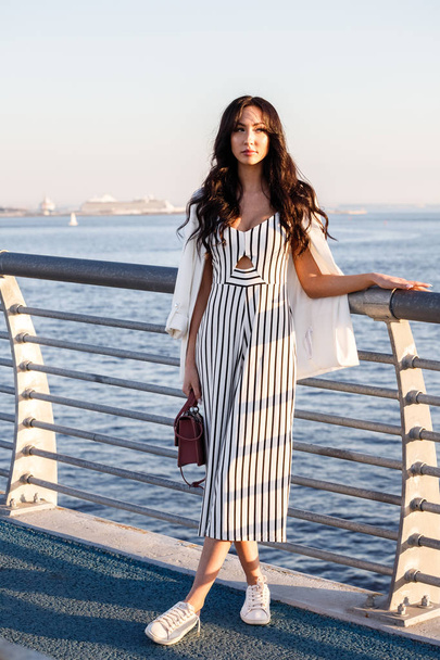 縞模様の白と黒のドレスで美しい若いブルネットの女性の屋外撮影。巻き毛の女性、日没時に桟橋に立って海の背景がぼやけてください。  - 写真・画像
