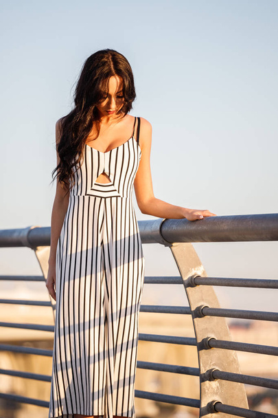 Outdoor shot of beautiful young brunette woman in striped black and white dress. Curly woman standing on pier during sunset, blurred sea background  - Fotografie, Obrázek