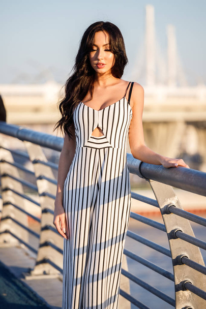 Outdoor shot of beautiful young brunette woman in striped black and white dress. Curly woman standing on pier during sunset, blurred sea background  - Zdjęcie, obraz