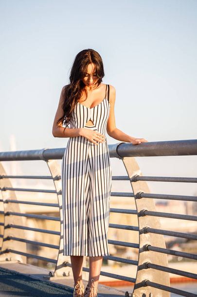 Outdoor shot of beautiful young brunette woman in striped black and white dress. Curly woman standing on pier during sunset, blurred sea background  - Φωτογραφία, εικόνα