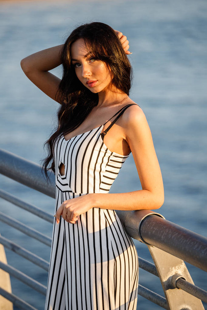 Outdoor shot of beautiful young brunette woman in striped black and white dress. Curly woman standing on pier during sunset, blurred sea background  - Fotoğraf, Görsel