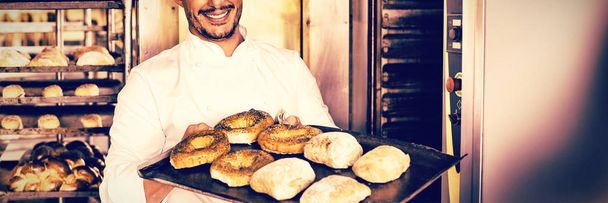 Happy baker showing tray of fresh bread in the kitchen of the bakery - Fotografie, Obrázek