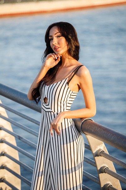 Outdoor shot of beautiful young brunette woman in striped black and white dress. Curly woman standing on pier during sunset, blurred sea background  - Фото, зображення