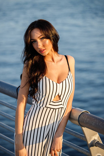Outdoor shot of beautiful young brunette woman in striped black and white dress. Curly woman standing on pier during sunset, blurred sea background  - Fotó, kép