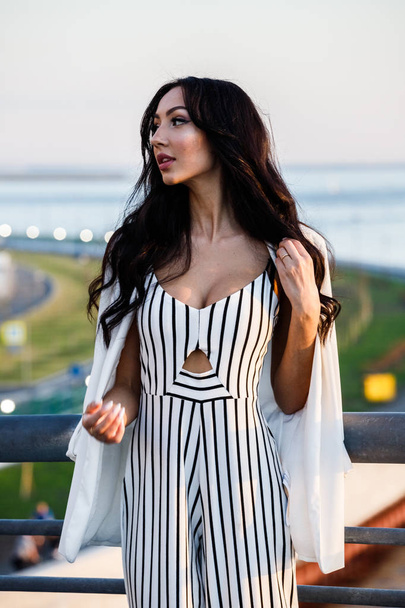 Outdoor shot of beautiful young brunette woman in striped black and white dress. Curly woman standing on city embankment in sunlight, blurred city view background  - Foto, Imagen