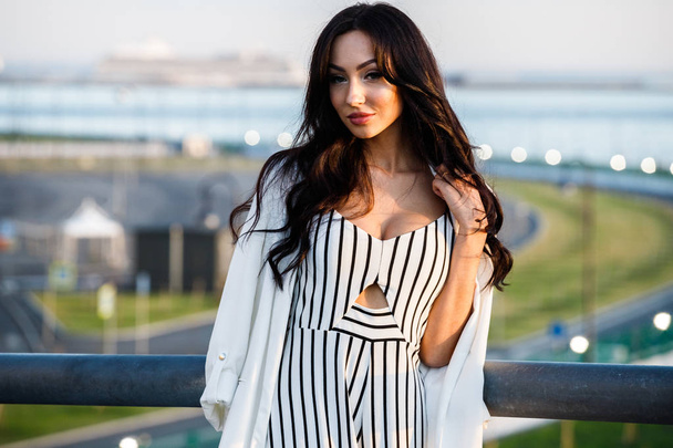 Outdoor shot of beautiful young brunette woman in striped black and white dress. Curly woman standing on city embankment in sunlight, blurred city view background  - Valokuva, kuva