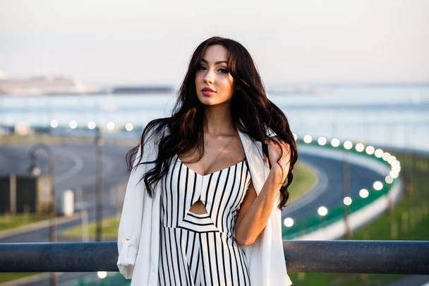 Outdoor shot of beautiful young brunette woman in striped black and white dress. Curly woman standing on city embankment in sunlight, blurred city view background  - Valokuva, kuva