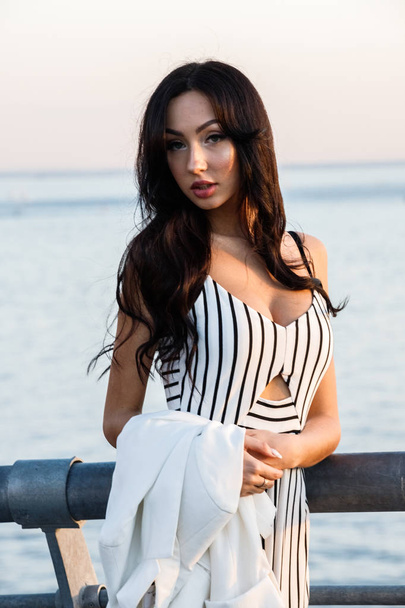 Outdoor shot of beautiful young brunette woman in striped black and white dress. Curly woman standing on pier during sunset, blurred sea background  - Foto, immagini