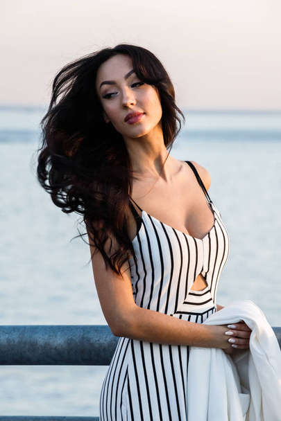 Outdoor shot of beautiful young brunette woman in striped black and white dress. Curly woman standing on pier during sunset, blurred sea background  - Photo, image