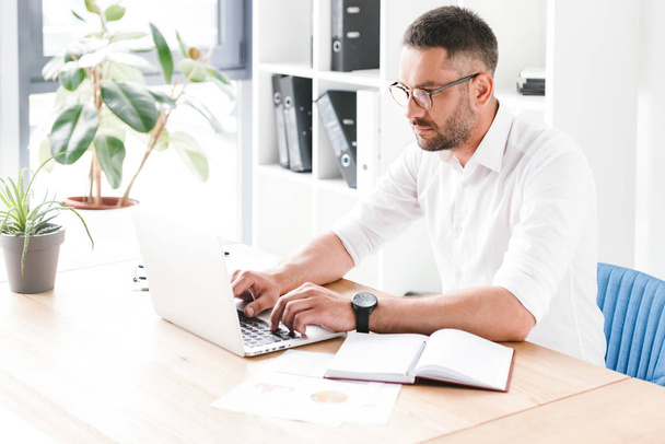 Photo of serious bearded man 30s in white shirt sitting at table and typing on laptop while working in bright office room - Foto, afbeelding