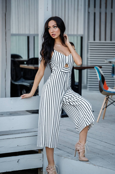 Outdoor portrait of beautiful young brunette woman in striped black and white dress. Curly woman standing in cafe interior - Φωτογραφία, εικόνα