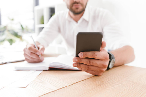 Cropped image of concentrated businessman in white shirt sitting at table and writing down information at notebook from his smartphone while working in bright office room - Foto, immagini
