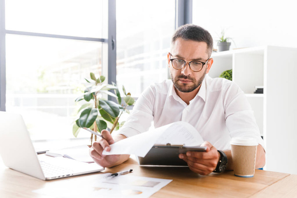 Portrait of serious businesslike man 30s in white shirt holding and examining paper documents during work in office - Foto, imagen
