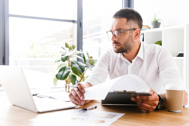 Portrait of smart businesslike man 30s in white shirt verifying information from paper documents using laptop during work in office - Foto, Imagem