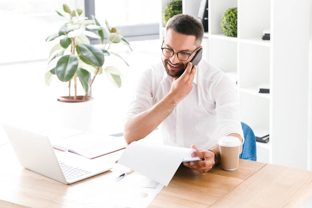 Portrait of happy businesslike man 30s in white shirt talking on cell phone and holding clipboard with paper documents while sitting at table in office - Foto, imagen