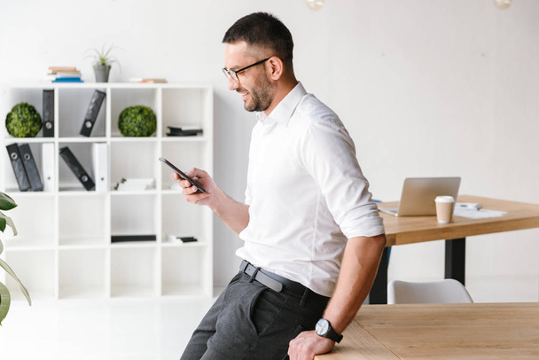 Красивый веселый мужчина в белой рубашке сидит на столе в офисе и использует смартфон для бизнес-чата
 - Фото, изображение