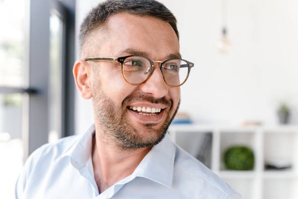 Portrait closeup of handsome man 30s wearing white shirt and glasses looking aside while standing near big window in business centre or office room - Фото, зображення