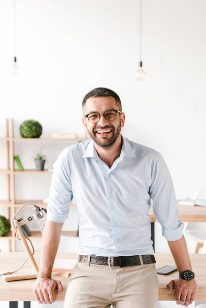 Happy portrait of businesslike man wearing white shirt sitting on table in office and smiling at camera - Фото, изображение