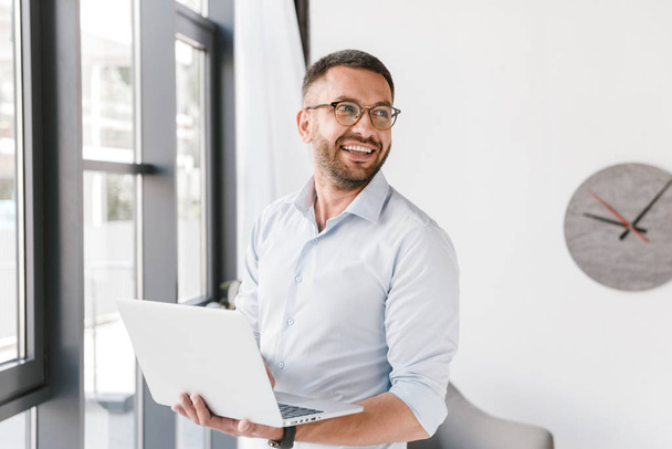 Image of joyful business man wearing white shirt looking aside at copyspace while holding silver laptop during work in office interior - Foto, afbeelding