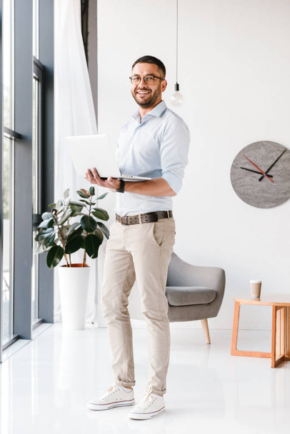 Full length photo of attractive modern man wearing eyeglasses looking at camera while standing and working on laptop in office - Фото, изображение