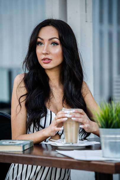 Ritratto di bella giovane donna bruna in abito bianco e nero a righe. donna riccia bere caffè e leggere libro in caffè interno
 - Foto, immagini