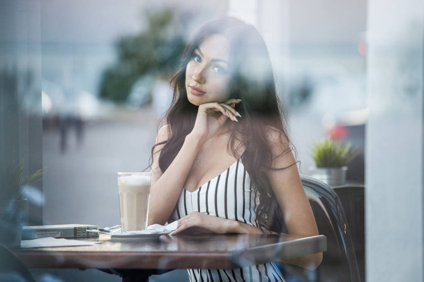 Retrato de una hermosa joven morena en vestido blanco y negro a rayas. Mujer rizada bebiendo café y leyendo libro en el interior de la cafetería
 - Foto, Imagen