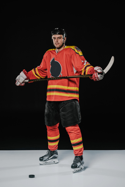 full length view of professional ice hockey player holding hockey stick and looking at camera on black  - Photo, Image