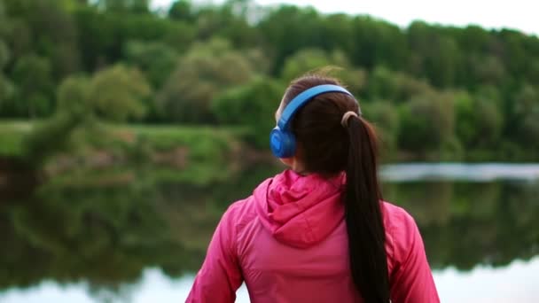A girl in a pink jacket and blue headphones stands with her back and looks at the river early in the morning after a run - Video, Çekim