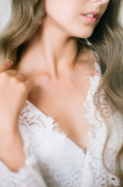 Beautiful young bride with long blond curly hair in a long white lacy dressing gown in the morning at the mirror - Fotoğraf, Görsel