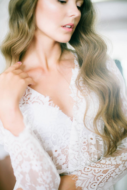 Beautiful young bride with long blond curly hair in a long white lacy dressing gown in the morning at the mirror - 写真・画像