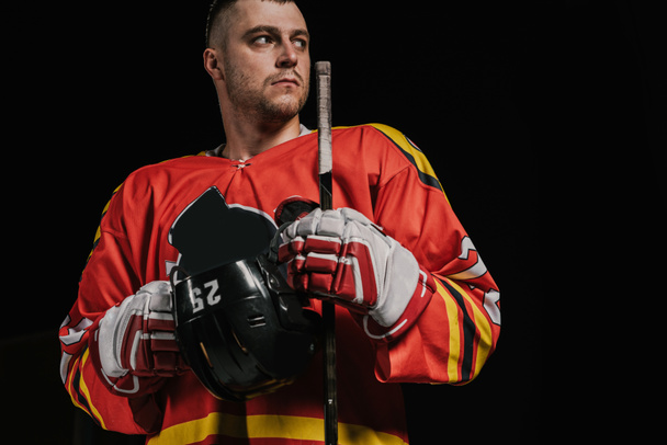 professional sportsman holding hockey stick with helmet and looking away isolated on black - Photo, Image