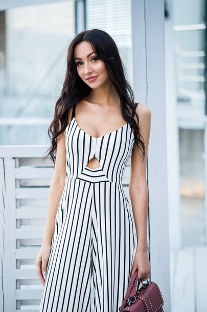 Outdoor portrait of beautiful young brunette woman in striped black and white dress. Curly woman standing in cafe interior - Photo, image