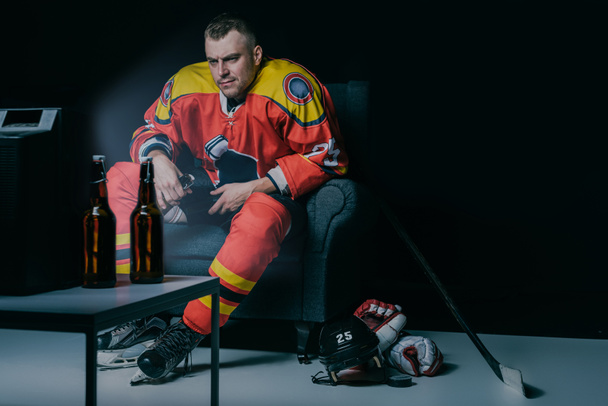 serious young hockey player drinking beer and watching tv while sitting in armchair in black - Photo, Image