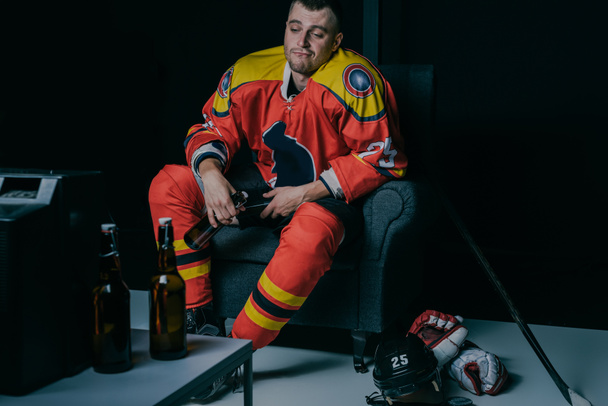 junger Eishockeyspieler trinkt Bier und schaut fern, während er in einem schwarzen Sessel sitzt - Foto, Bild