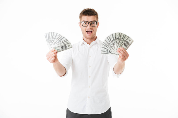 Portrait of an excited young man in eyeglasses holding money banknotes isolated over white background - Photo, Image