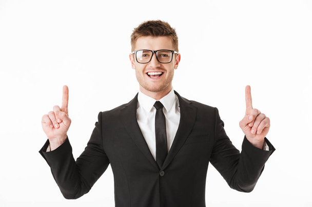 Portrait of a happy young businessman in suit and eyewear pointing fingers up at copy space isolated over white background - Φωτογραφία, εικόνα