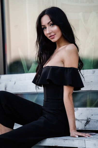 Portrait of beautiful young brunette woman in black dress with stylish makeup sitting on wooden bench, green plants background - Фото, изображение
