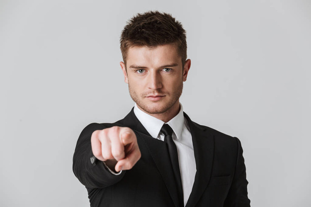 Portrait of a confident young businessman in suit pointing finger at camera isolated over gray background - Fotografie, Obrázek