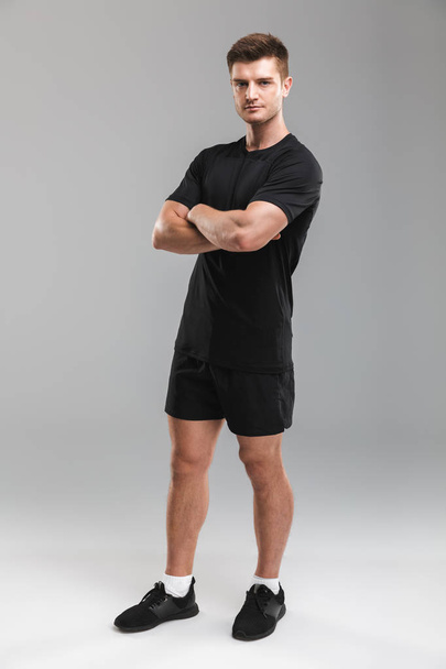 Full length portrait of a confident young sportsman standing with arms folded isolated over gray background - Foto, Imagen