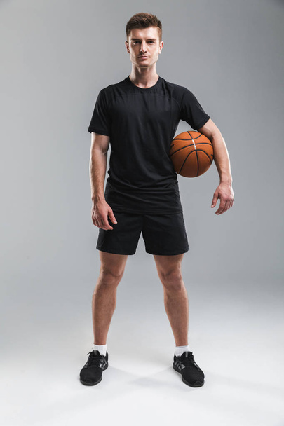 Full length portrait of a concentrated young sportsman standing with a basketball isolated over gray background - Foto, imagen