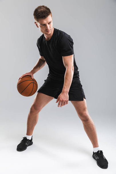 Full length portrait of a confident young sportsman playing basketball isolated over gray background - 写真・画像