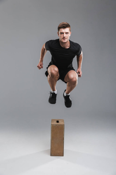 Full length portrait of a concentrated young sportsman jumping over a wooden box isolated over gray background - Foto, imagen