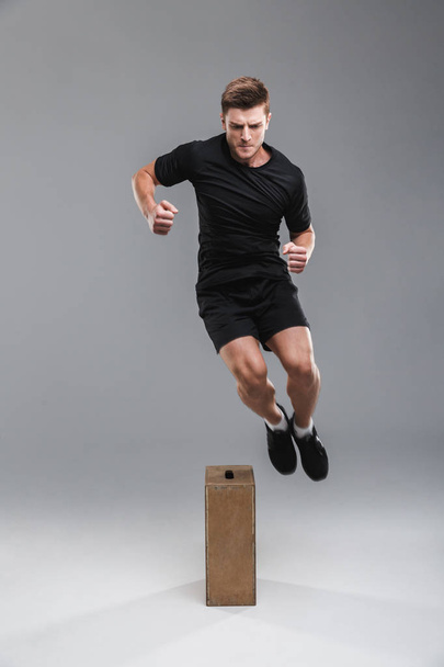 Full length portrait of a motivated young sportsman jumping over a wooden box isolated over gray background - Foto, Imagem