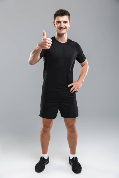 Full length portrait of a smiling young sportsman showing thumbs up isolated over gray background - 写真・画像